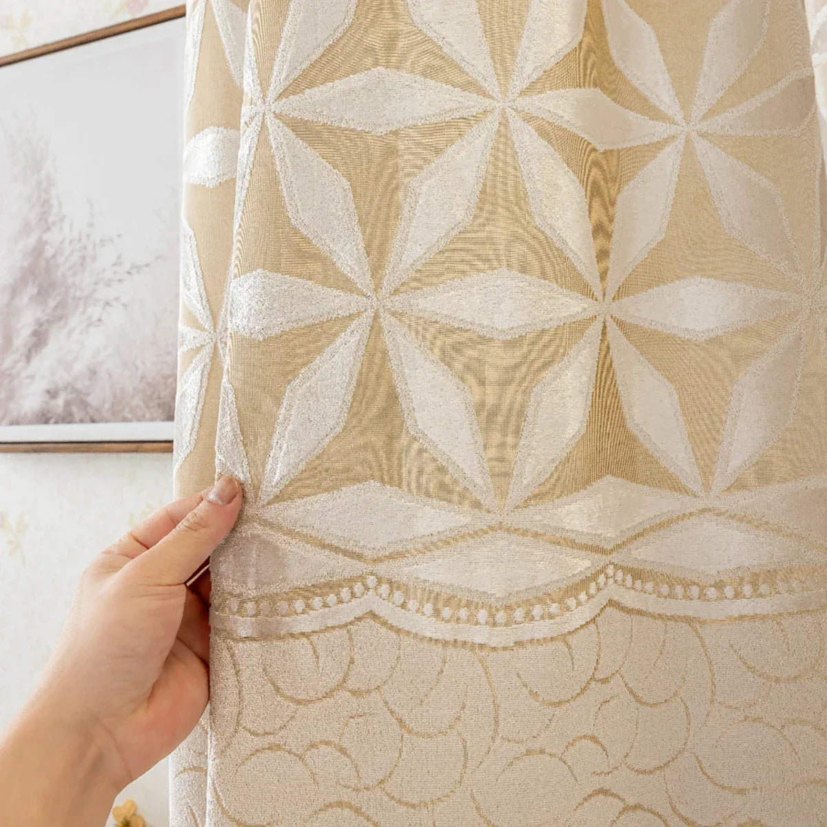 Hand holding beige curtain fabric showing white geometric flowers above a scalloped embroidered border.