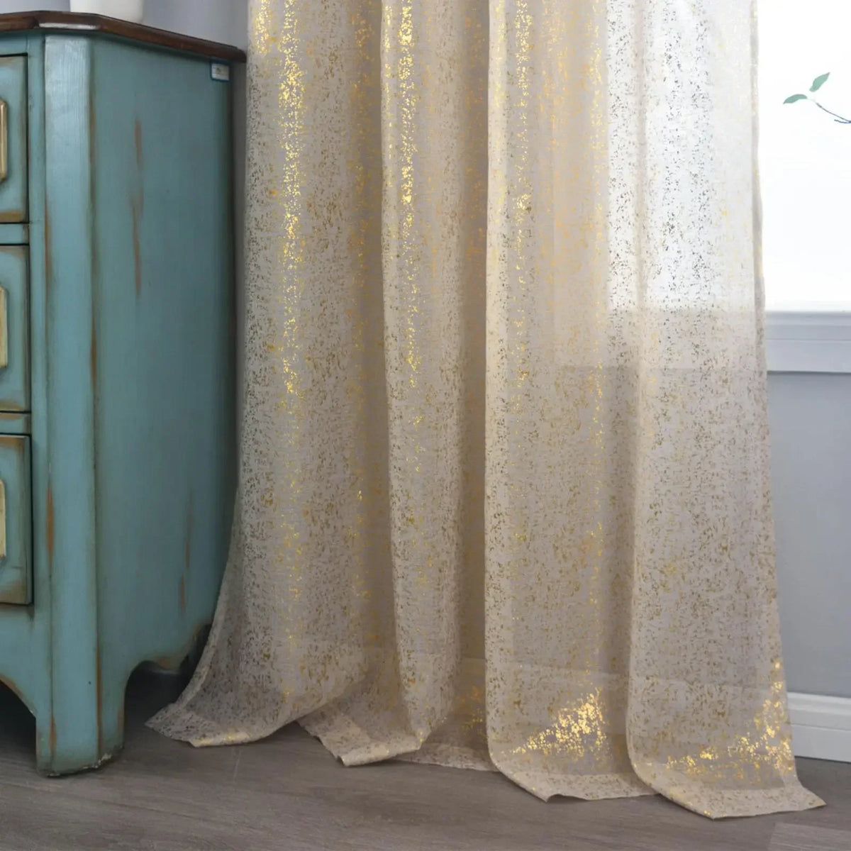 Beige sheer curtains with gold-coloured speckles pooling slightly on the floor beside a blue-green cabinet.