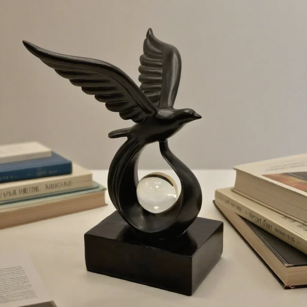 Black bird sculpture with raised wings and a clear sphere on a black base beside books.