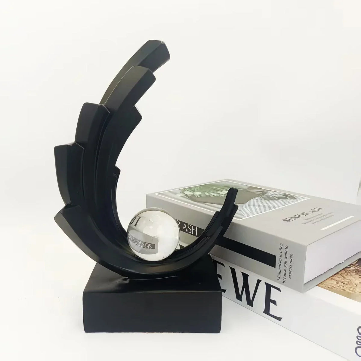Black stepped crescent sculpture with a clear sphere on a rectangular base beside books.