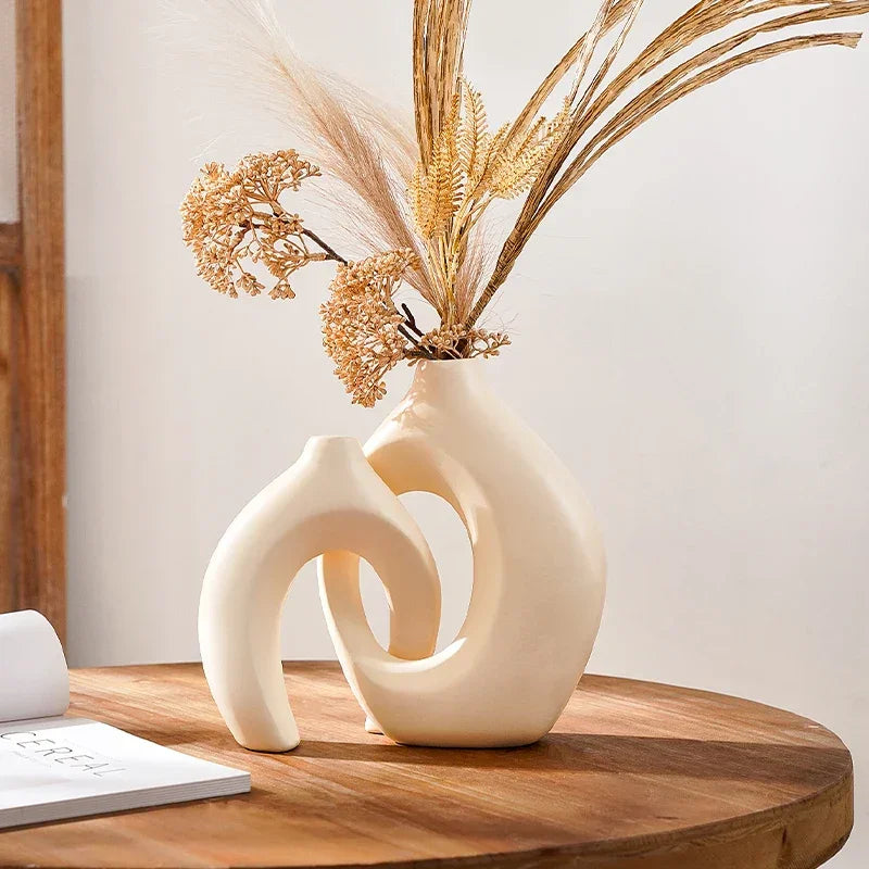 Cream sculptural vases with interlocking open centres, displayed with dried stems on a round wooden table