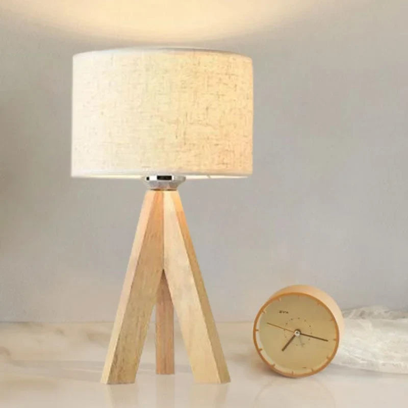 Wooden tripod table lamp with an illuminated cream shade beside a small round clock.