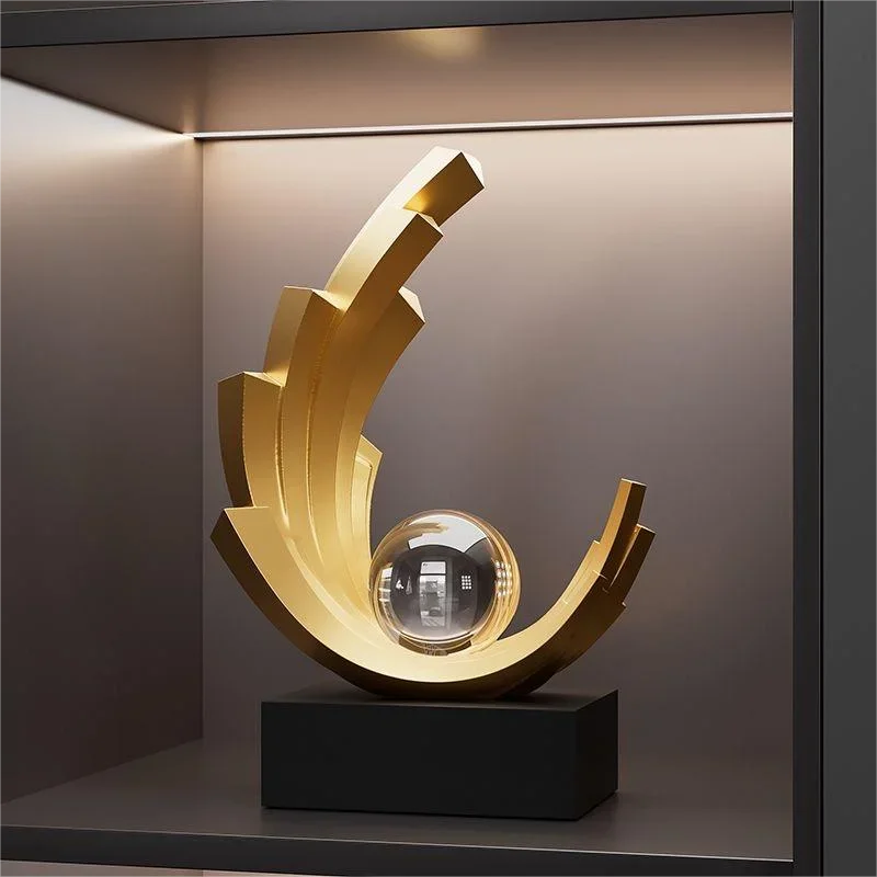 Gold-coloured abstract crescent sculpture with stepped edges and a clear sphere on a black rectangular base.