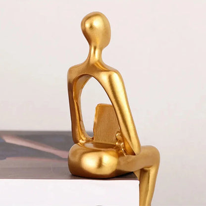 Gold-coloured abstract seated figure holding a book, with its legs hanging over the edge of a book