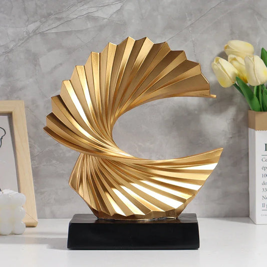 Gold-coloured abstract wave sculpture with pleated folds and an open centre on a black rectangular base.