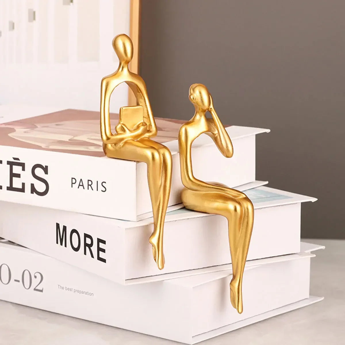 Two gold-coloured seated figures displayed on stacked books, one holding a book and one resting its face on a hand