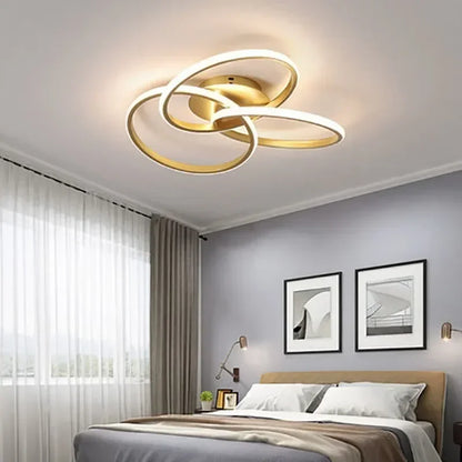 Gold-coloured trefoil LED ceiling light above a bed in a grey room with framed artwork and a large window