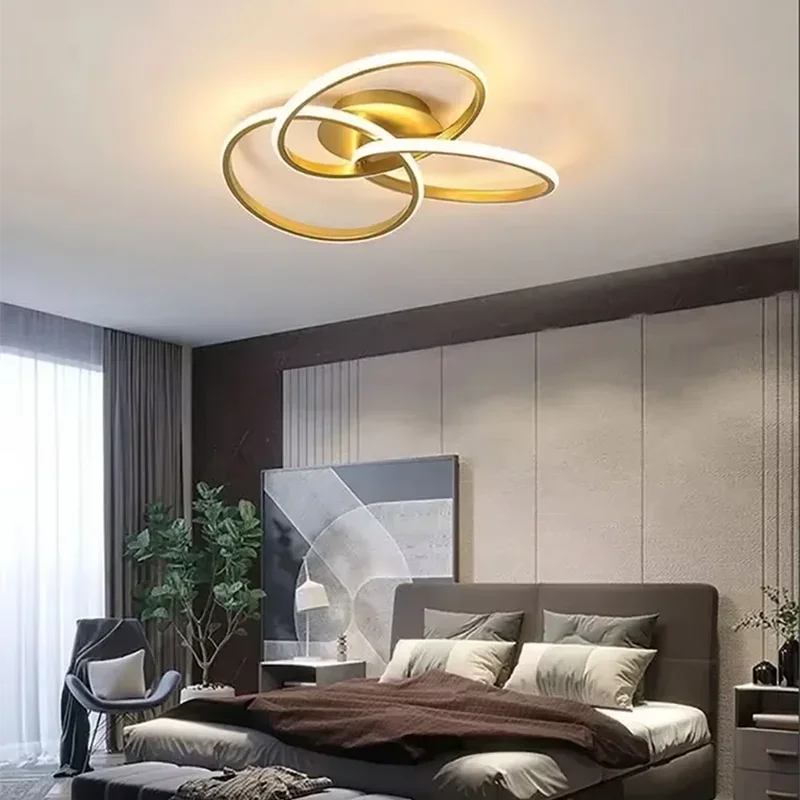 Gold-coloured trefoil LED ceiling light with three intertwined illuminated loops above a dark upholstered bed