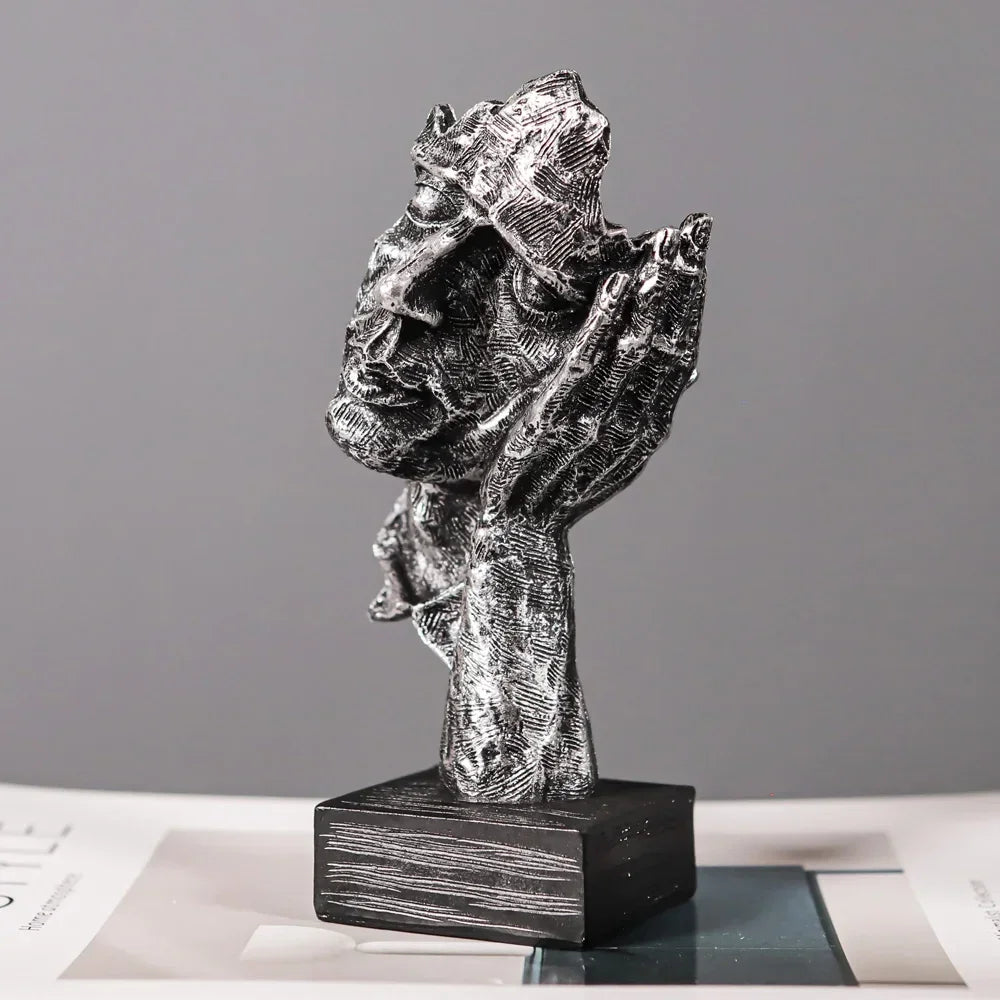 Angled view of a silver-coloured face sculpture with a hand beside its ear and a black rectangular base