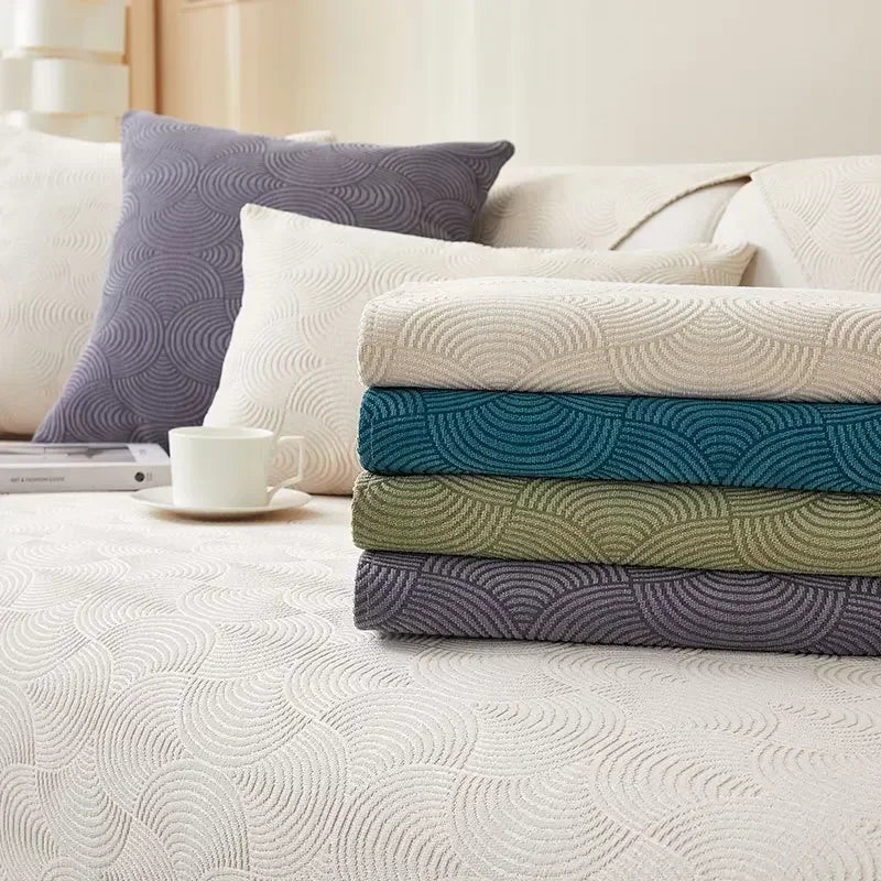 Folded sofa covers in cream, blue, green and grey-purple stacked on a pale covered sofa.