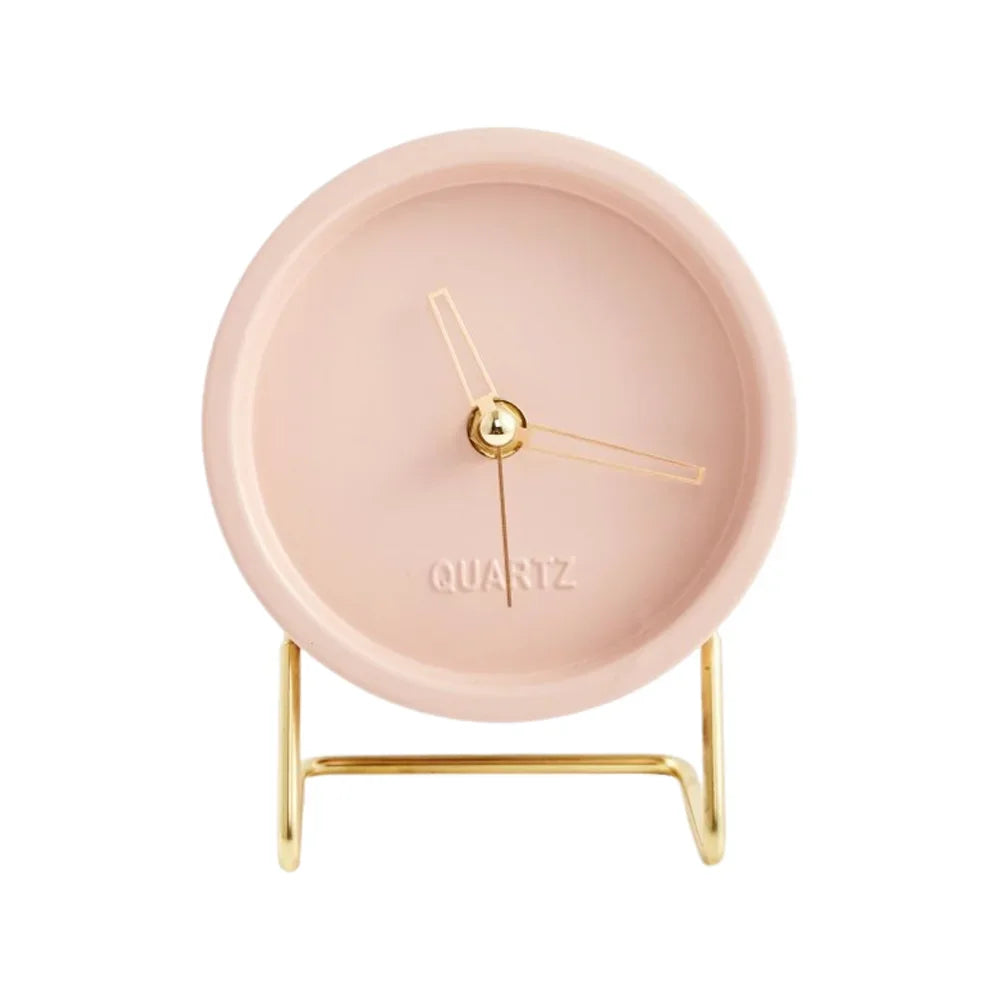 Pink round desk clock with gold-coloured hands and an angular wire stand on a white background