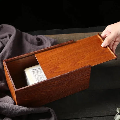 A hand sliding the panel of a rectangular wooden tissue box cover open to reveal a tissue pack.