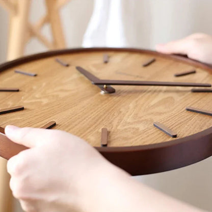 Hands holding a round wooden wall clock, showing its wood-grain face and raised dark hour markers