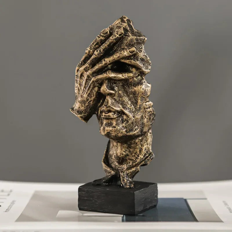 Gold-coloured textured face sculpture with a hand covering its eyes, mounted on a black rectangular base