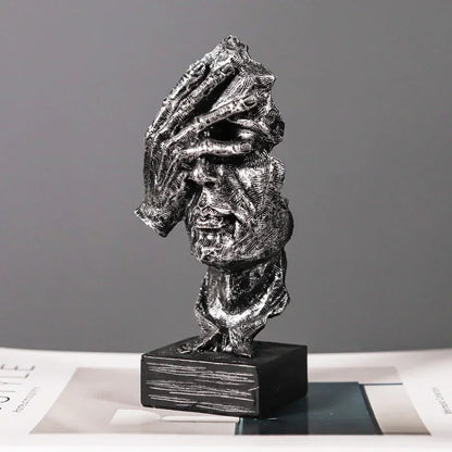 Silver-coloured textured face sculpture with a hand covering its eyes, mounted on a black base