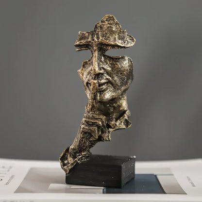 Gold-coloured fragmented face sculpture with a finger held to its lips, mounted on a black rectangular base
