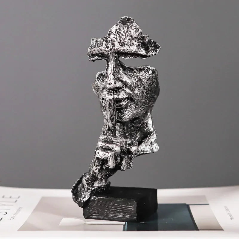 Silver-coloured fragmented face sculpture with a finger held to its lips, mounted on a black base