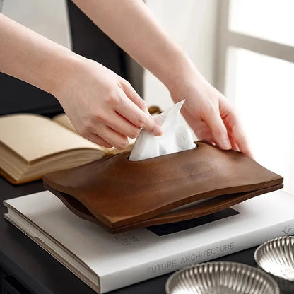 Close view of hands removing a tissue from a curved walnut wood tissue box cover placed on books.